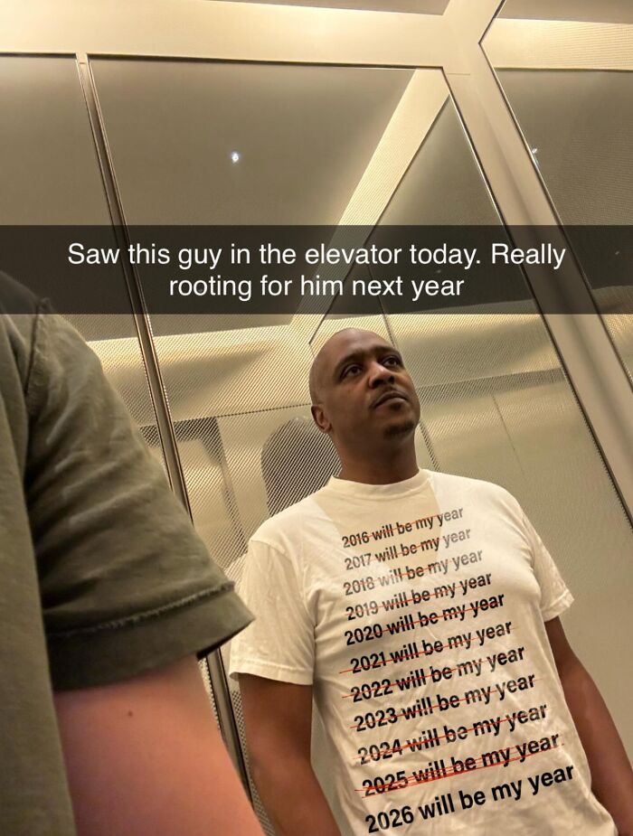 A man in an elevator wearing a white T-shirt with a list of years, 2016-2026, many crossed out saying, will be my year.