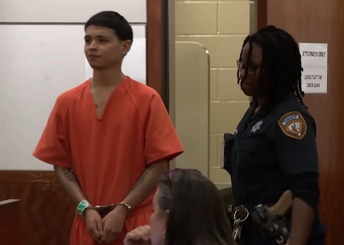 Man in orange prison uniform and handcuffs stands with female officer in courtroom Man in orange prison uniform and handcuffs stands with female officer in courtroom