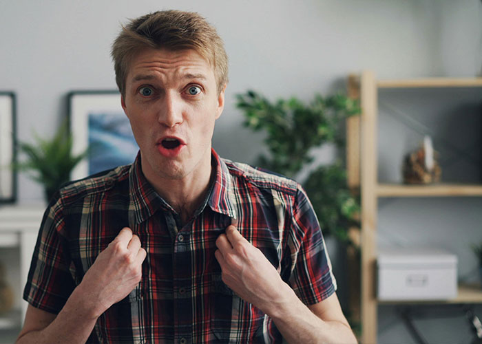 Man in a plaid shirt with wide eyes and an open mouth, pointing to himself in disbelief, often linked to low intelligence.