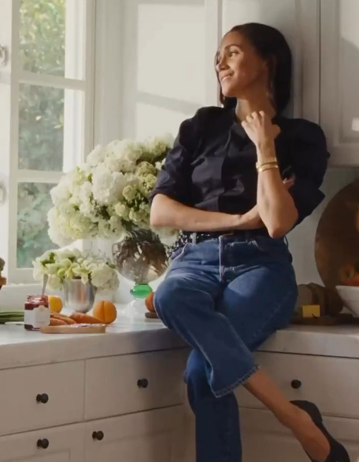 Meghan Markle, wearing a dark top and jeans, sits on a kitchen counter, smiling and looking out a window. Meghan Markle, wearing a dark top and jeans, sits on a kitchen counter, smiling and looking out a window.