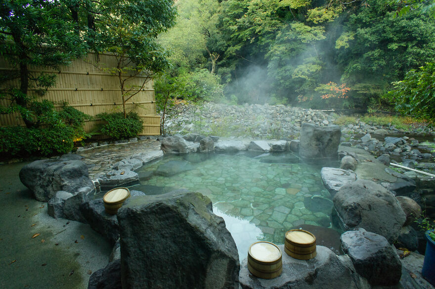 A steaming Japanese hot spring, or onsen, surrounded by large rocks and lush green trees. Two wooden buckets sit on the rocks.