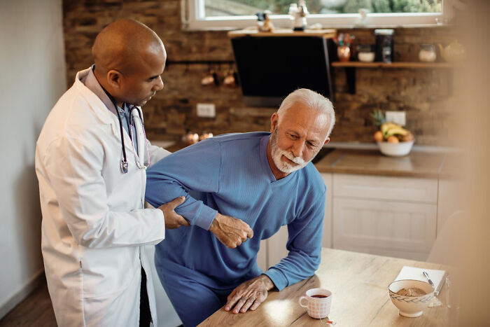 A doctor in a white coat assists an elderly man in blue pajamas, helping him stand up from a kitchen table. They cross to the other side.