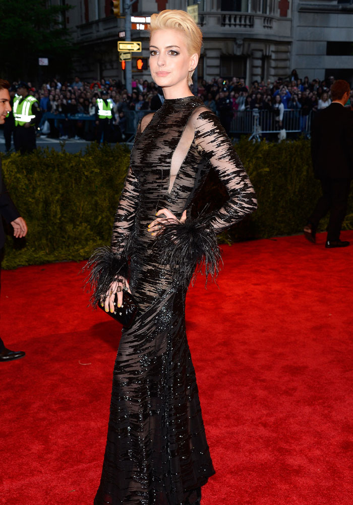 Anne Hathaway in a bold sheer outfit, wearing a black sequined and feathered gown with a blonde pixie cut on the red carpet.