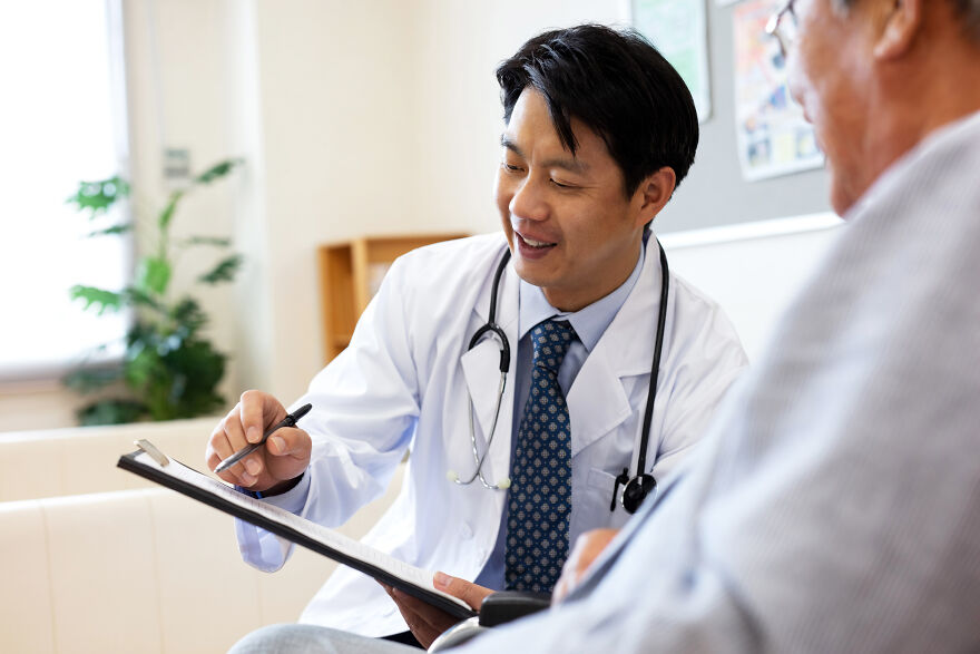 A smiling Japanese doctor in a white coat with a stethoscope, explaining health information to a patient.