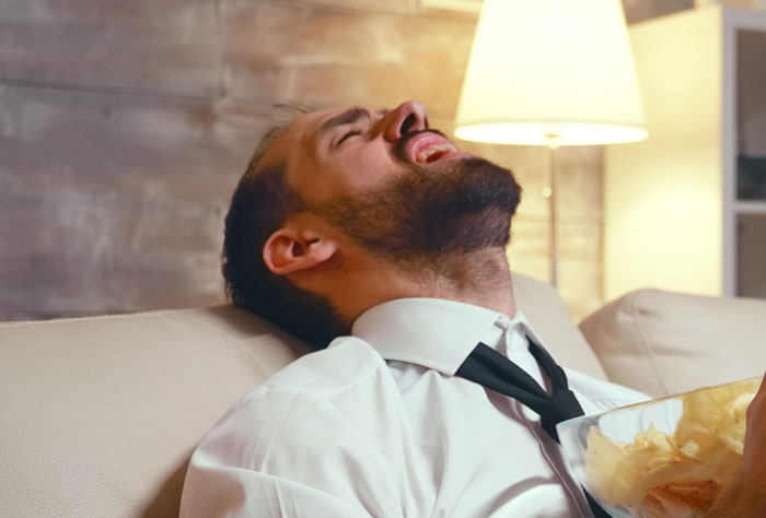 Man in white shirt laughing on sofa with bowl of chips