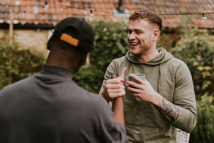 Two men chatting outdoors, one laughing while holding a mug. The image shows the dynamics of celebrity relatives.