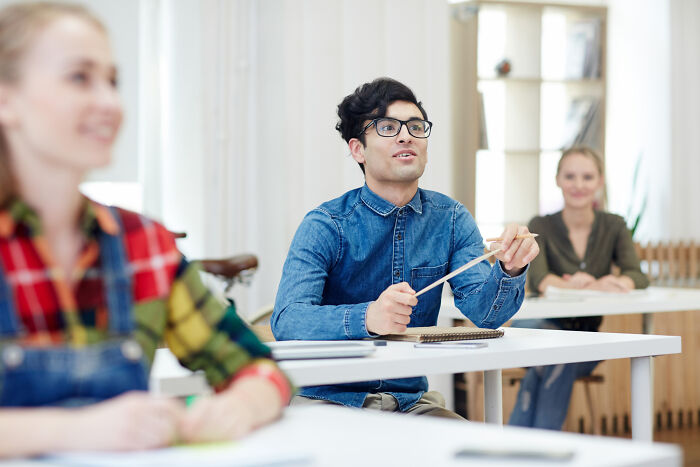 A young man in glasses and a denim shirt holds pencils, looking upward, while other students attend a class, depicting second-hand embarrassment.