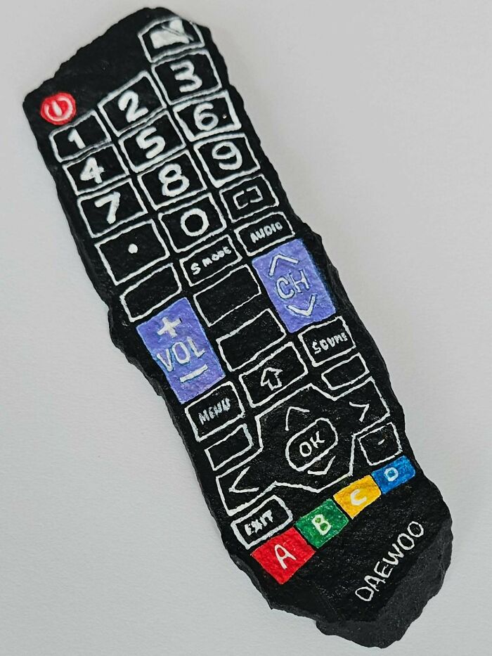 A black rock painted as a Daewoo TV remote control. This stone creation features white button outlines and colorful details.