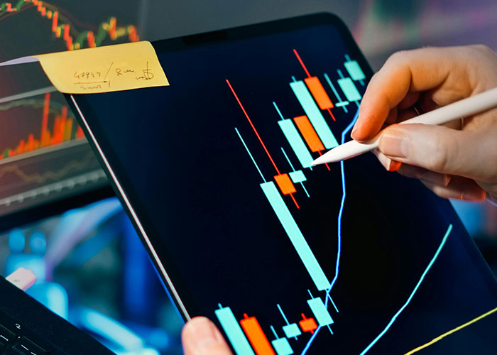 A hand uses a stylus on a tablet displaying stock market data. A sticky note is attached to the screen. People of low intelligence might struggle with interpreting such complex financial charts.