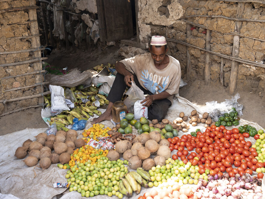 Man selling diverse fresh produce like coconuts, tomatoes, and limes, highlighting varying diets globally.