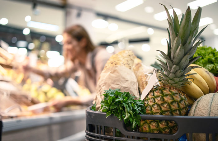 A shopping cart filled with groceries like pineapple, bread, and greens. A blurry woman shops in the background. Wrong facts on Reddit can be misleading, especially about nutrition.