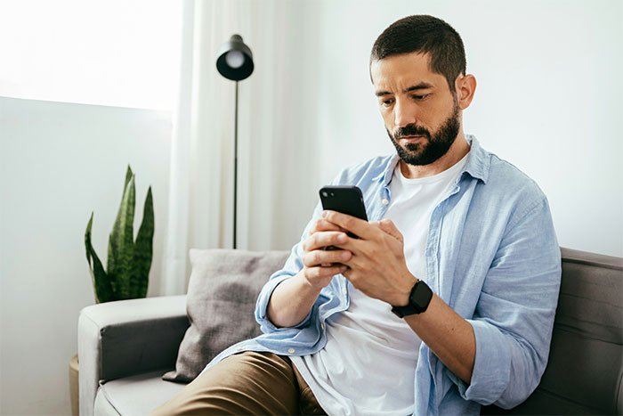 A man with a beard sits on a sofa looking intently at his phone, reacting to shocking grandchild news.