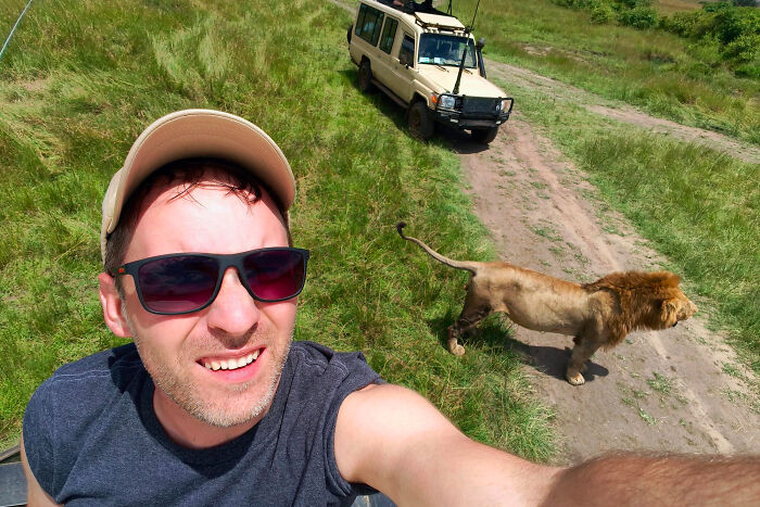 A man taking a selfie with a lion and safari vehicle in the background. Tourists often do annoying things abroad.