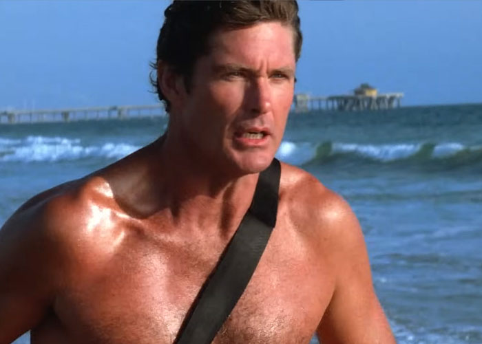 A shirtless lifeguard with a concerned expression, on a beach with ocean waves and a pier in the background. US stereotypes.