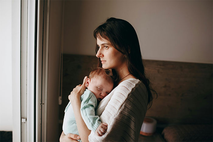 A thoughtful young mother holds her sleeping baby, contemplating family dynamics and the unhinged MIL situation.
