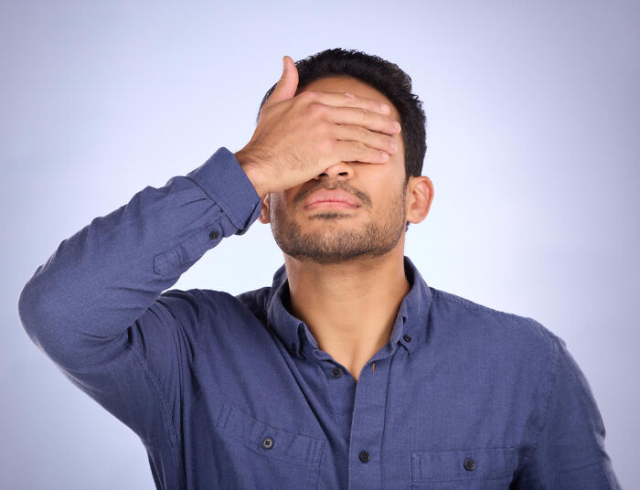 A man in a blue shirt covering his eyes with his hand, conveying embarrassment or disbelief, common feelings in hilarious engagement stories.