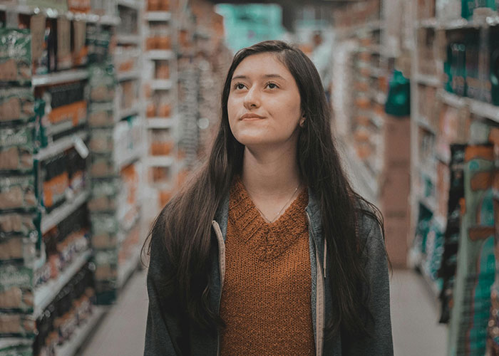 A young woman looking up thoughtfully in a grocery aisle, pondering the economy and her wallet.