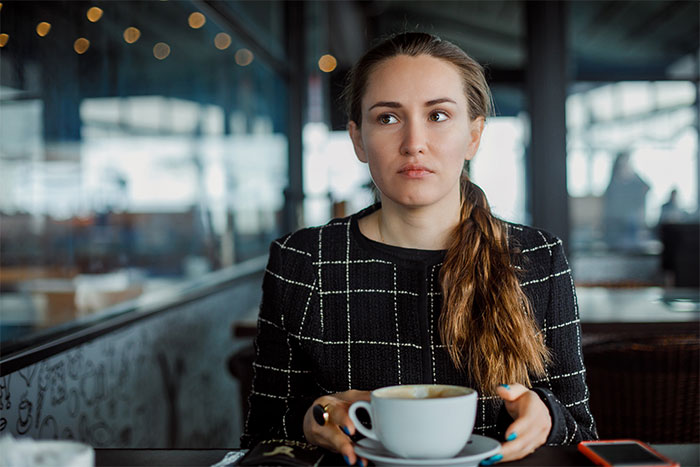 A woman with a thoughtful expression in a black plaid jacket holds a coffee cup, contemplating family dynamics and grandchild care.