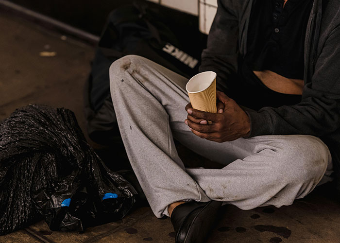 A person sits on the ground, holding an empty paper cup, next to a black bag. This image debunks stereotypes about the US.