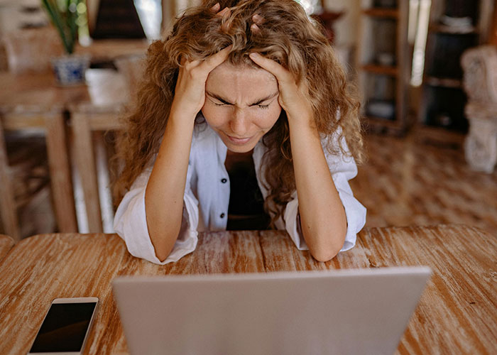 A woman with curly hair looks frustrated, holding her head, worrying about the economy and her wallet while using a laptop.