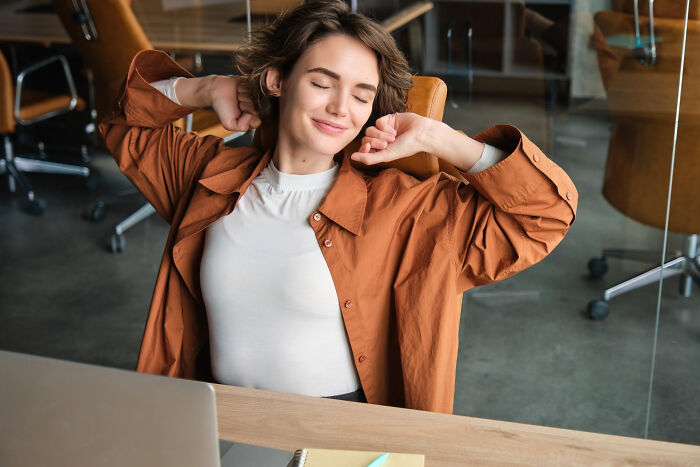 A young woman, eyes closed, stretching in an office chair. She is an employee relaxing between savage things to do.