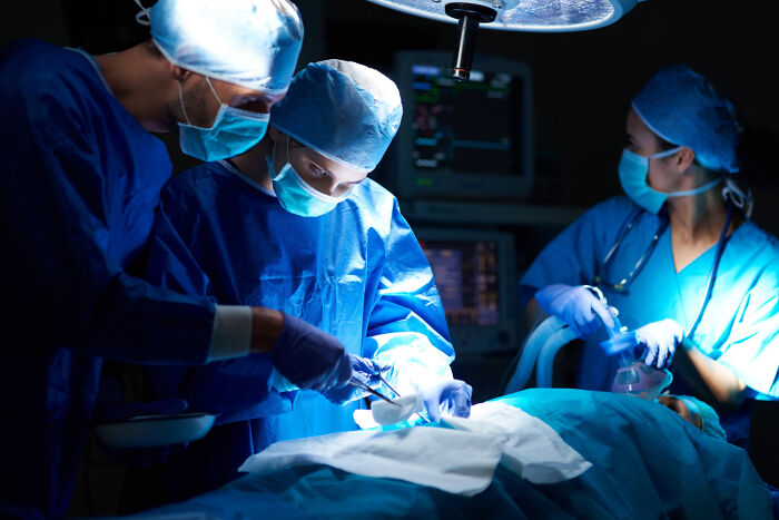 Medical team performing surgery. One surgeon holds an instrument while another tends to the patient, evoking "cross to the other side" stories.