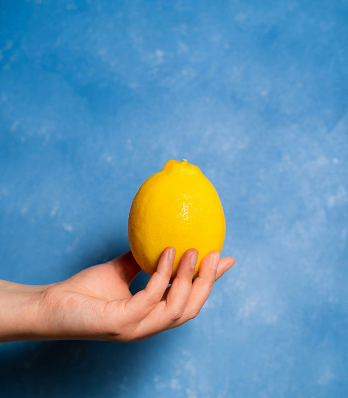 A hand holding a single yellow lemon against a blue background, a weird package, possibly delivered by mailmen.