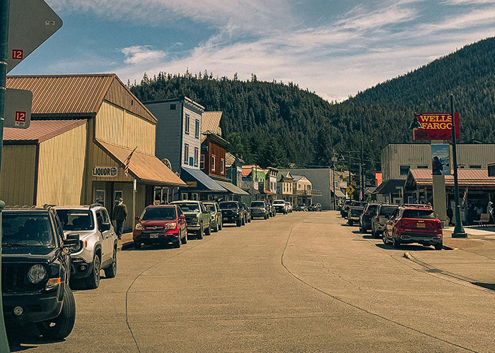 A street in a small US town, lined with cars and buildings, backed by a lush green mountain. Stereotypes about the US.