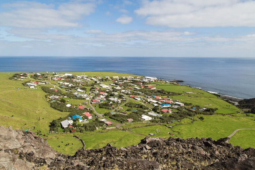 An aerial view of an isolated village nestled on a green coast, with the vast blue ocean stretching to the horizon.