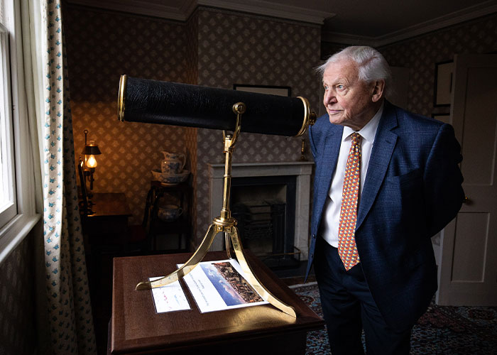 David Attenborough in a suit and tie, looking out a window next to a telescope. His long life secrets are inspiring.