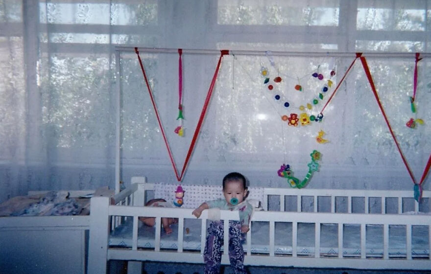 A baby with a pacifier stands in a crib with colorful decorations, symbolizing the demanding and stressful jobs of parenthood.