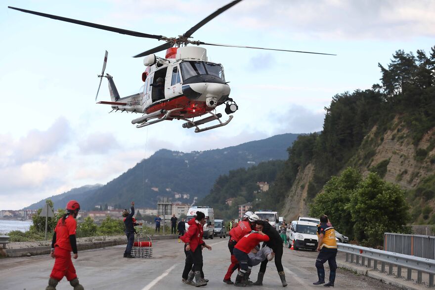 A rescue helicopter hovers over a road where emergency workers in red uniforms assist someone, highlighting stressful jobs.