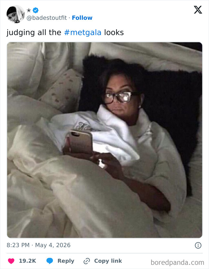 A person in bed, wearing glasses and a robe, looking intently at their phone. MET Gala 2026 reactions.