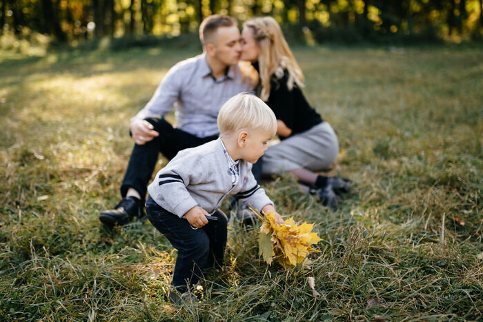 A toddler gathering yellow leaves in a grassy park, while his parents share a loving kiss in the background, showing engagement stories.