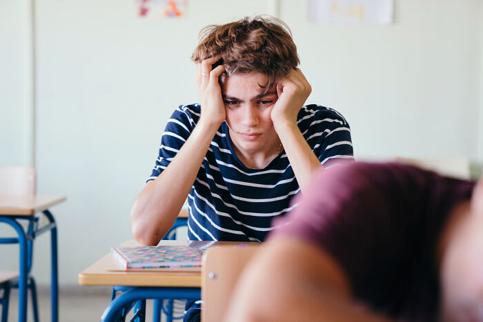 A young student with brown hair clutches their head, experiencing second-hand embarrassment, in a brightly lit classroom.