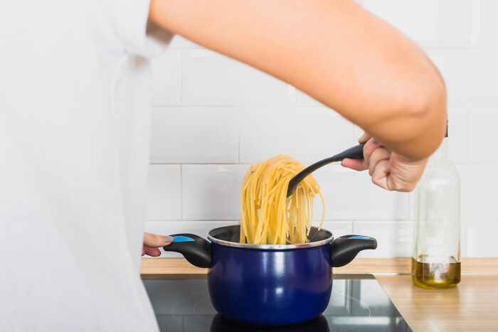 A person wearing a white shirt holds a spatula of spaghetti over a blue pot on an electric stove, symbolizing life's chaos.