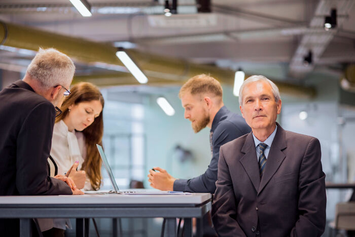 A businessman in a suit looks at the camera, with colleagues working in the background, sharing celebrity relatives stories.