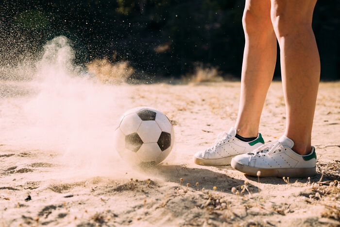 Person kicking a soccer ball on dusty ground in casual sneakers outdoor