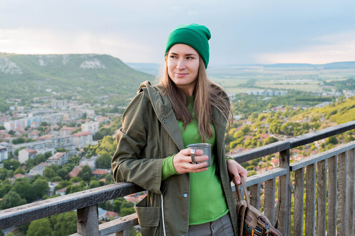 A woman in a green beanie and jacket stands on a balcony, holding a mug, overlooking a scenic town. Tourists behaving badly.