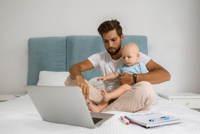A focused dad holds a baby while looking at a laptop, embodying balancing life and work. Savage things employees did.