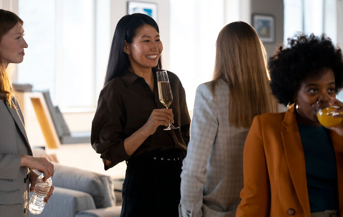 Diverse women conversing at a social event, one smiling with champagne. People watching can cause second-hand embarrassment.