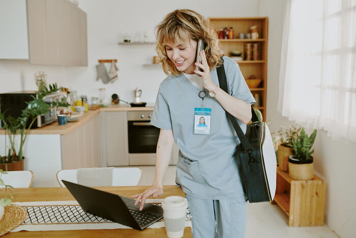 A smiling young employee in scrubs is on the phone while using a laptop in a kitchen, discussing savage things employees did.