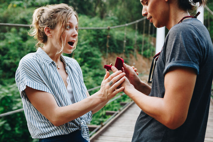 A man proposes to a surprised woman on a bridge in nature, showcasing romantic engagement stories.