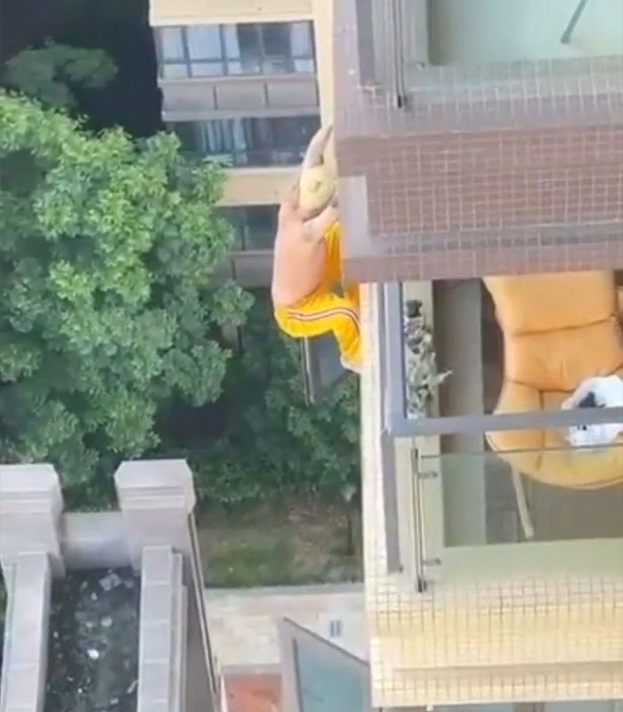 Man climbing rooftop ledge in yellow pants above apartment balcony