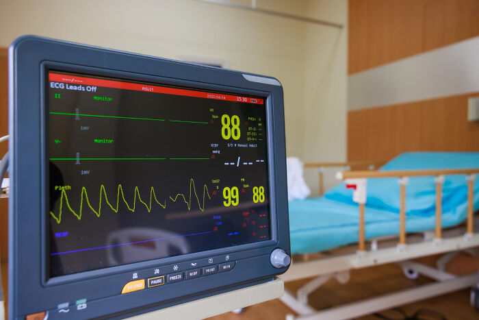 A hospital patient monitor displaying vital signs with a blurred empty bed in the background. Stories that will stay with you.