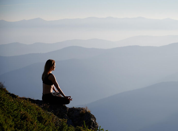 Woman meditating at sunset on a mountain cliff, silhouetted against hazy peaks. Stories that will stay with you.