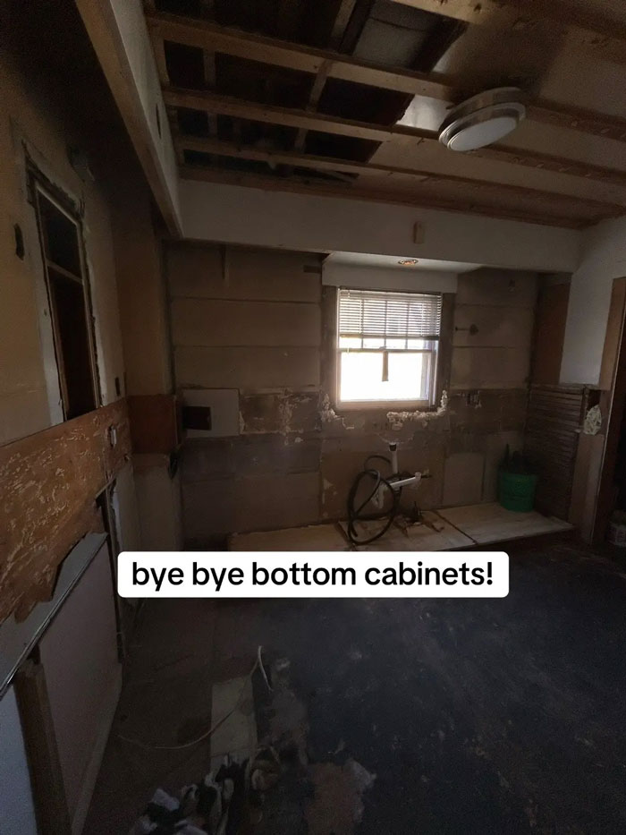 A kitchen under renovation with walls stripped, exposed ceiling beams, and a window. Sign says: bye bye bottom cabinets! An expensive contractor left the home.