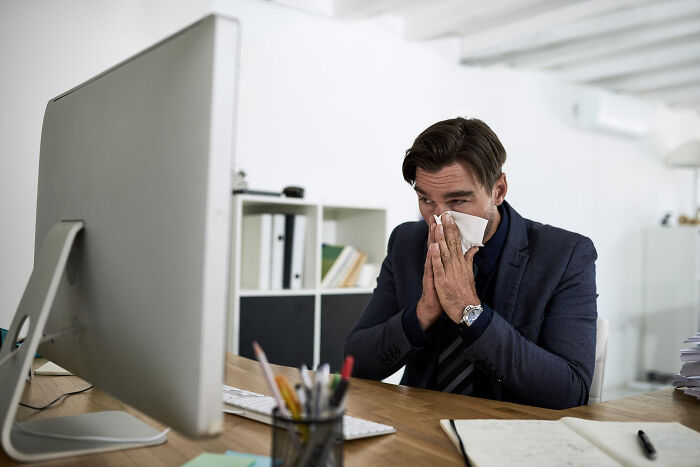 A man in a suit blows his nose with a tissue at his office desk, causing second-hand embarrassment.