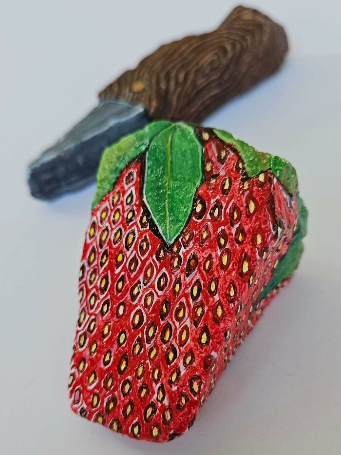 A vibrant strawberry rock creation with a painted green stem, showcasing colorful stone creations. A brown handle tool lies behind it.
