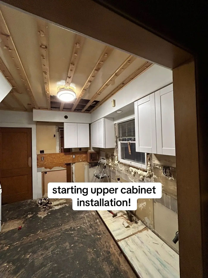 A kitchen under construction with exposed ceiling beams and newly installed upper cabinets, highlighting a contractor's work.